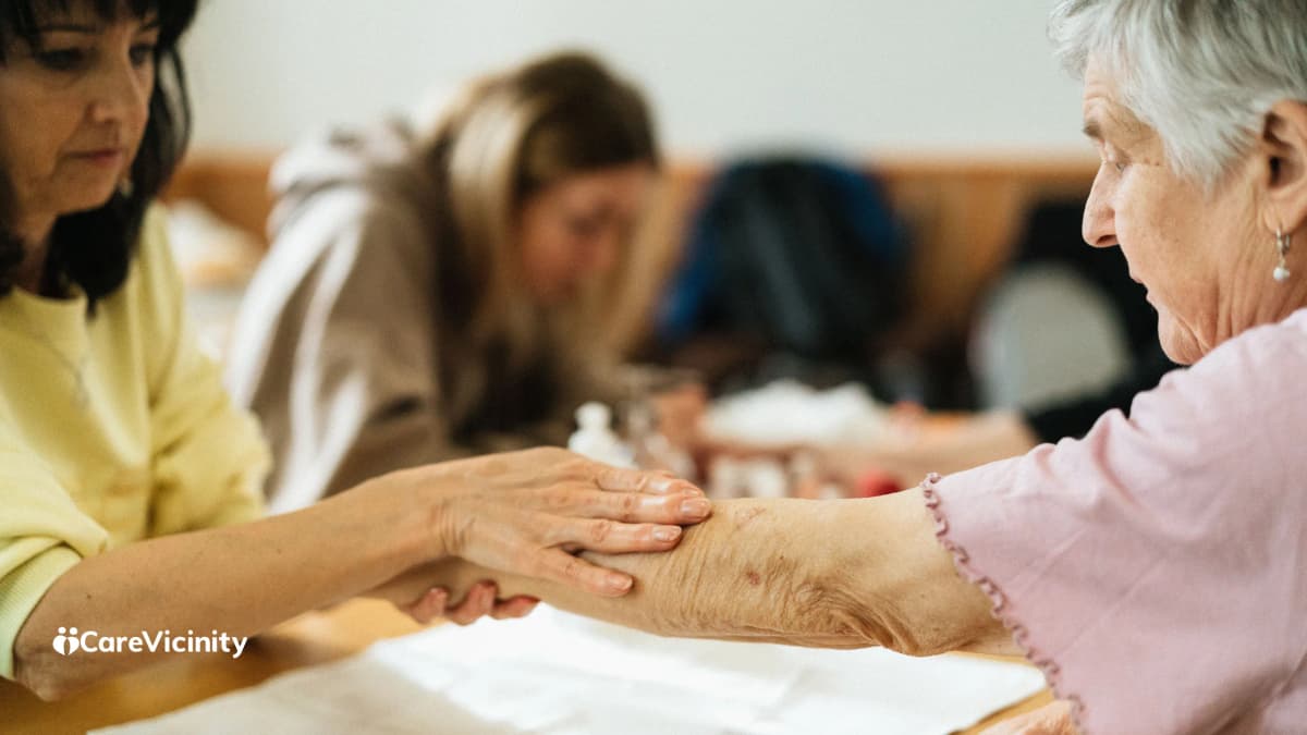 A support worker gently holding an older woman's forearm while seated together at a table