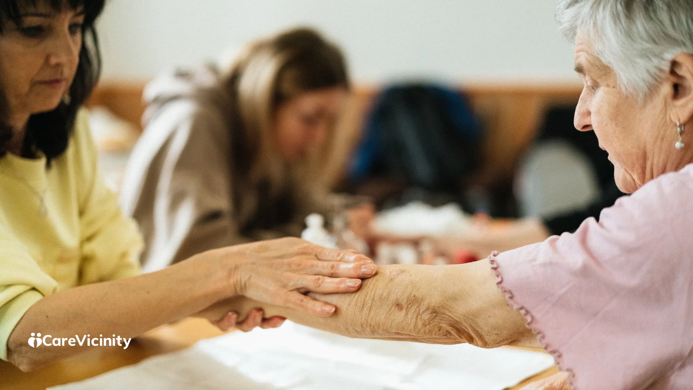 A support worker gently holding an older woman's forearm while seated together at a table