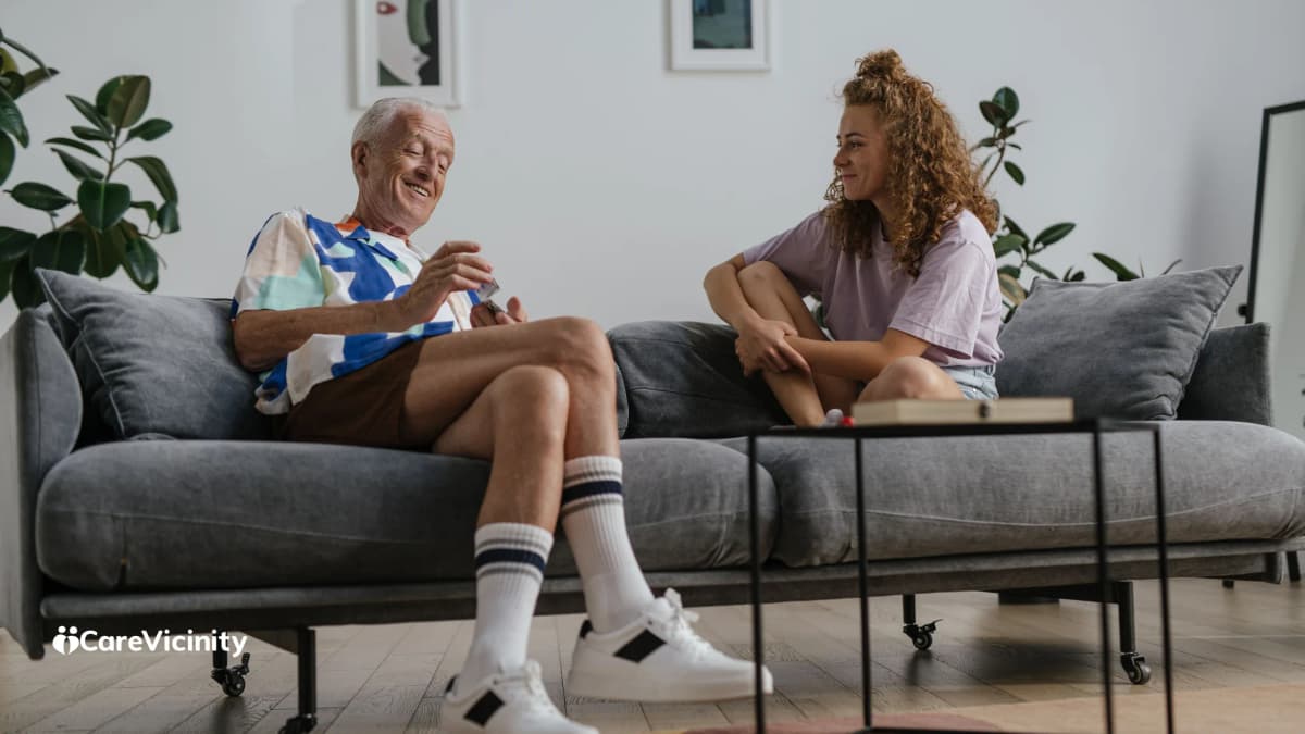 An older man and a younger support worker sitting together in a home living room, chatting warmly