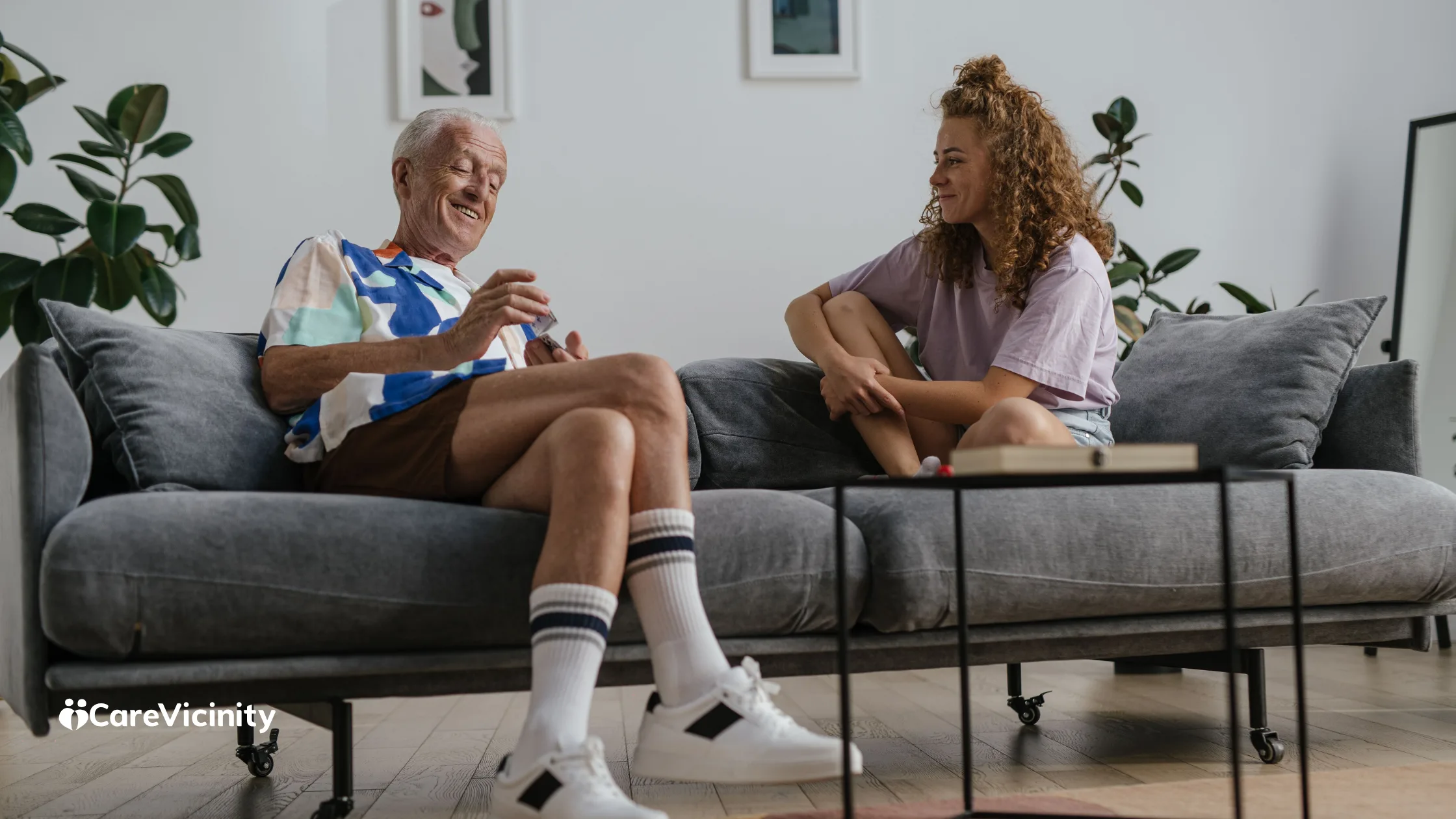 An older man and a younger support worker sitting together in a home living room, chatting warmly